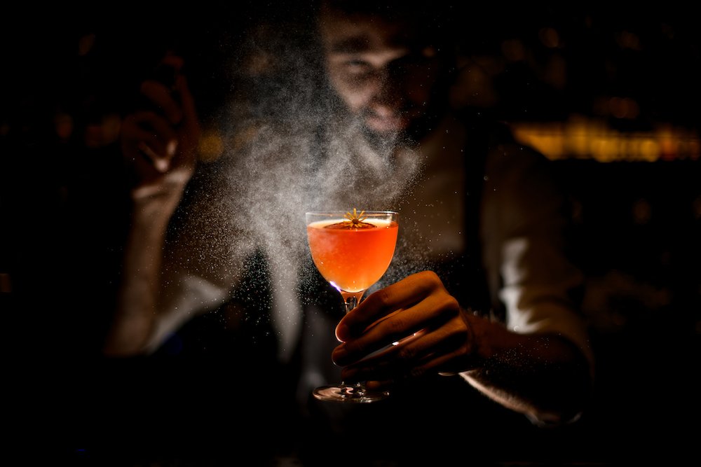 Professional bartender serving a cocktail with a lemon slice and a little yellow flower spraying on it in the dark on the bar counter